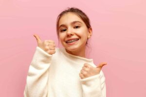 cheerful teenage girl with braces smiles and shows likes with her hands on a pink isolated background, child shows thumbs up and recommends