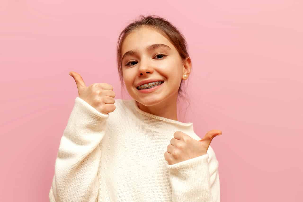 teenage girl with braces smiles and shows likes with her hands on pink isolated background, child shows thumbs up and recommends cheerful teenage girl with braces smiles and shows likes with her hands on a pink isolated background, child shows thumbs up and recommends