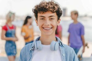 A portrait of a smiling teenage boy with curly hair and braces, wearing headphones and a blue shirt. He is standing outside in a city with friends, looking directly at the camera. The image captures a positive and authentic moment of youth and friendship.